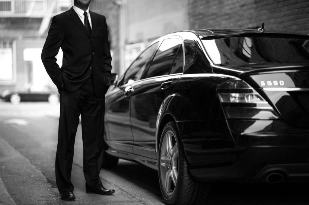 chauffeur privé en costume en attente aux cote de sa mercedes S 550 en noir et blanc